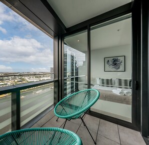 Un balcón con dos sillas verdes, una cama con decoración gris y blanca, y vista a la ciudad.