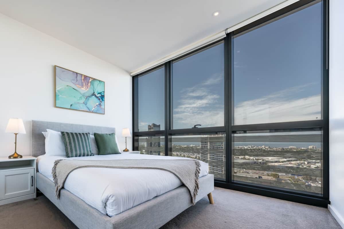 Ein modernes Schlafzimmer mit einem großen Bett, einem Nachttisch, einer Lampe, einem Gemälde und Blick auf die Stadt durch große Fenster.