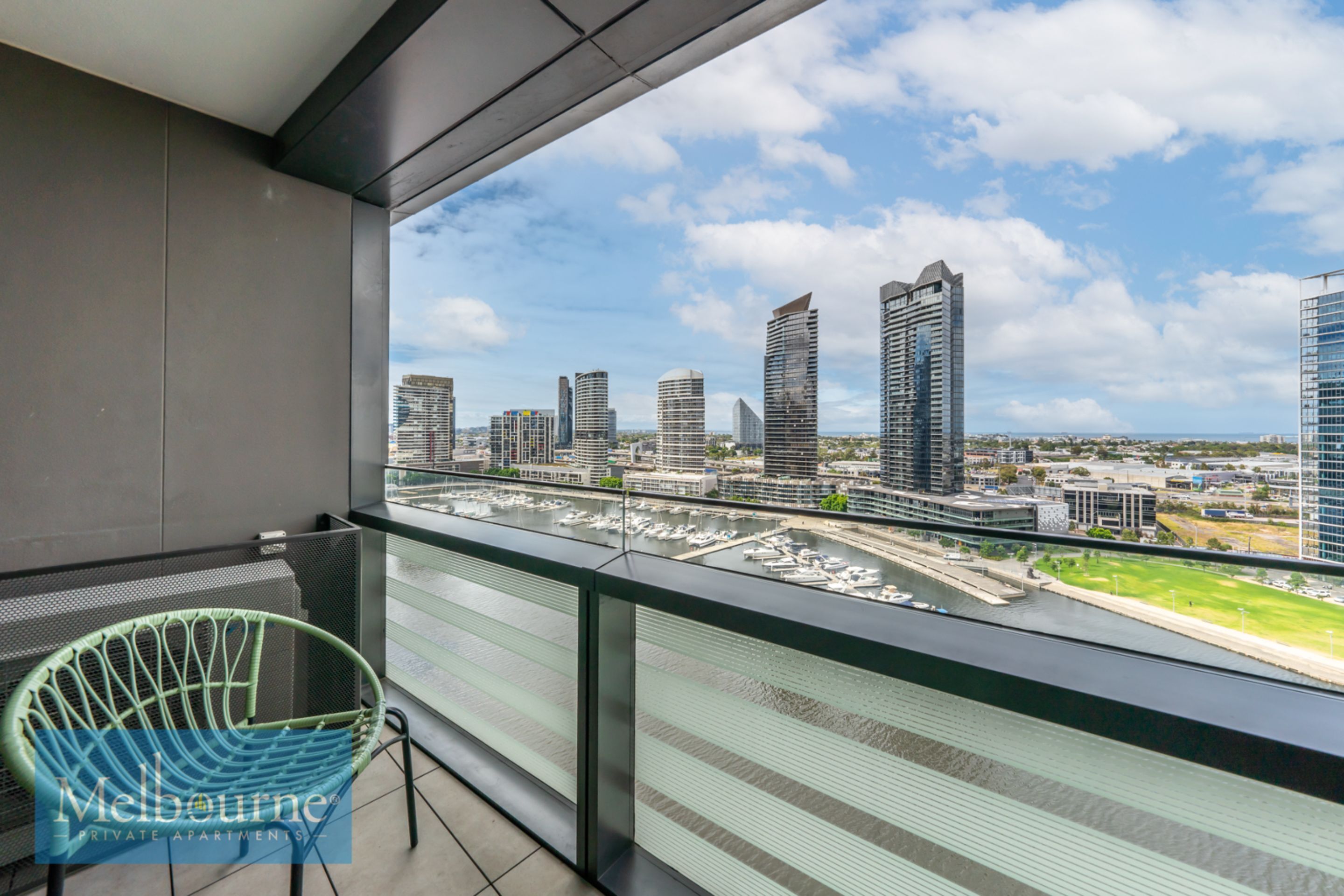 Un balcón con vistas a un horizonte urbano, una silla verde y una baranda de vidrio.