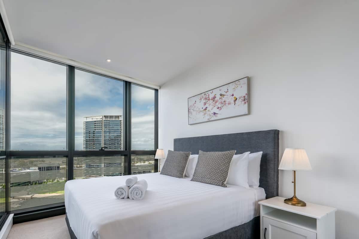 Ein modernes Hotelzimmer mit einem großen Bett, einem Nachttisch, einer Lampe und Blick auf die Stadt durch raumhohe Fenster.