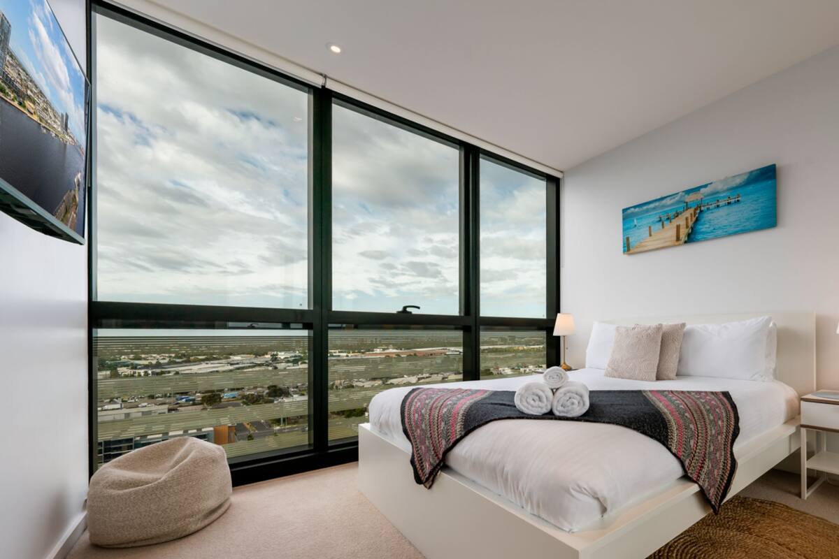Ein modernes Hotelzimmer mit großem Fenster und Blick auf die Stadt, einem ordentlich bezogenen Bett mit Kissen, einem Nachttisch mit Lampe sowie einem Bild an der Wand.