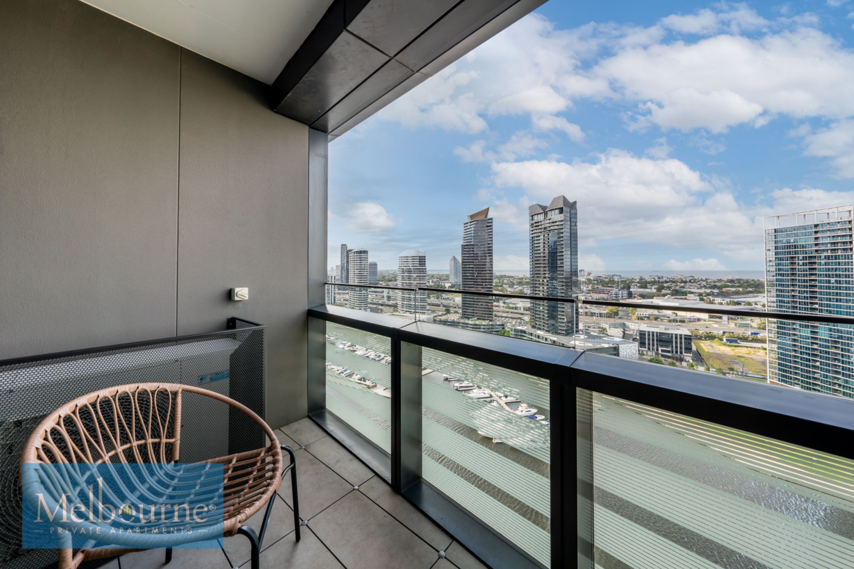 A balcony with a view of a cityscape, a wicker chair, and a glass railing.
