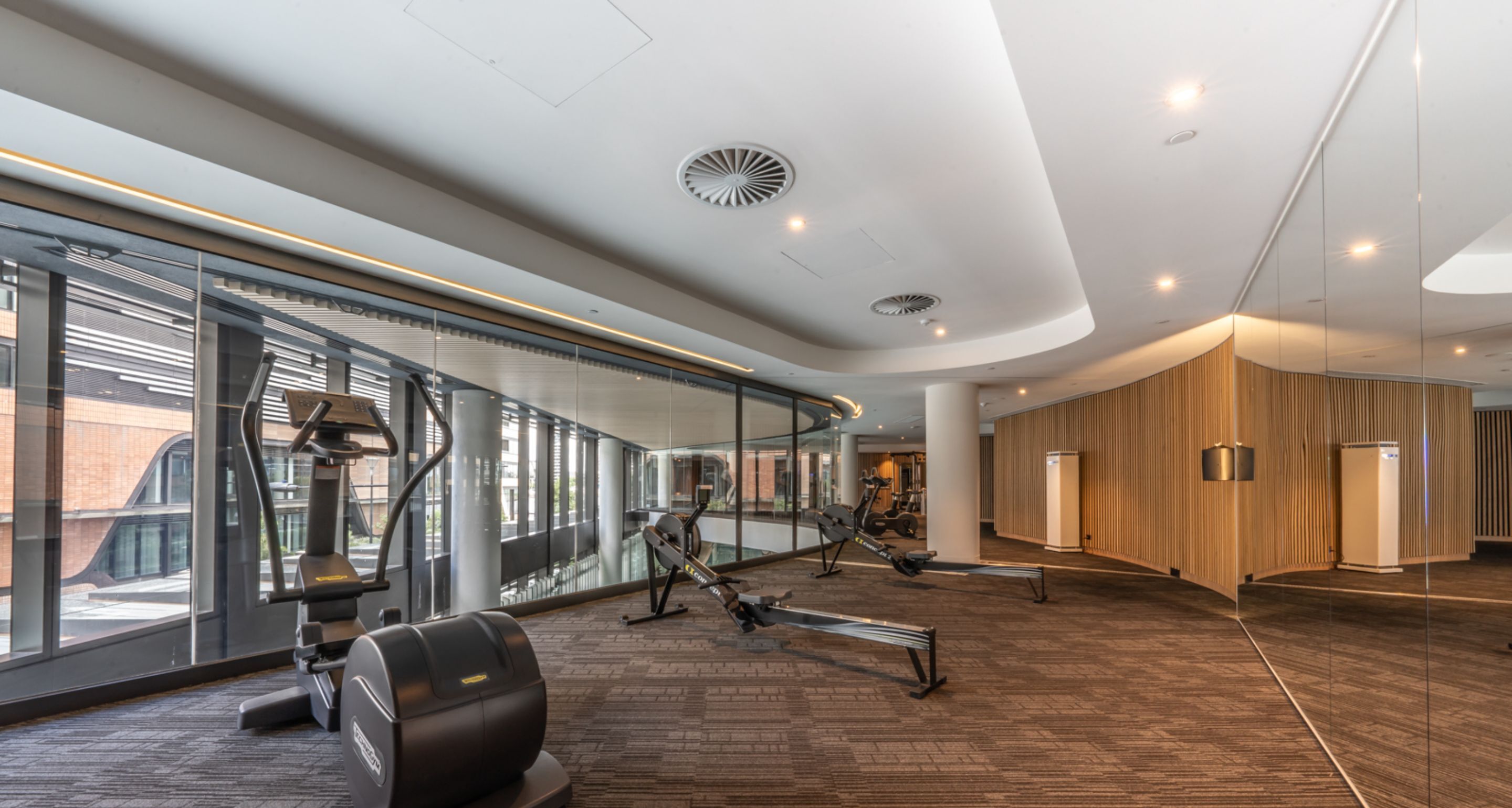 A modern gym with cardio machines, a wooden wall, and large windows.