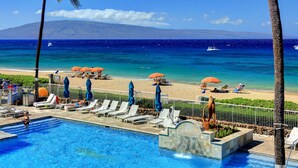 A pool area with lounge chairs and umbrellas, a beach with umbrellas, and a view of the ocean and a mountain in the background.