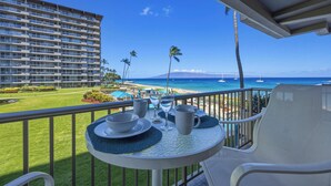 A balcony with a table set for two, overlooking a beach with palm trees and a hotel building in the background.