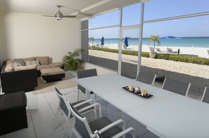 A modern room with a large window overlooking a beach, a white dining table with chairs, a sofa, and a ceiling fan. - Prime Residence #7 on 7 Mile Beach, High-End Ground Floor Beachfront Condo (George Town)