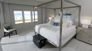 Un lit à baldaquin donnant sur la mer, une table de chevet avec des lampes et une valise posée sur le sol.