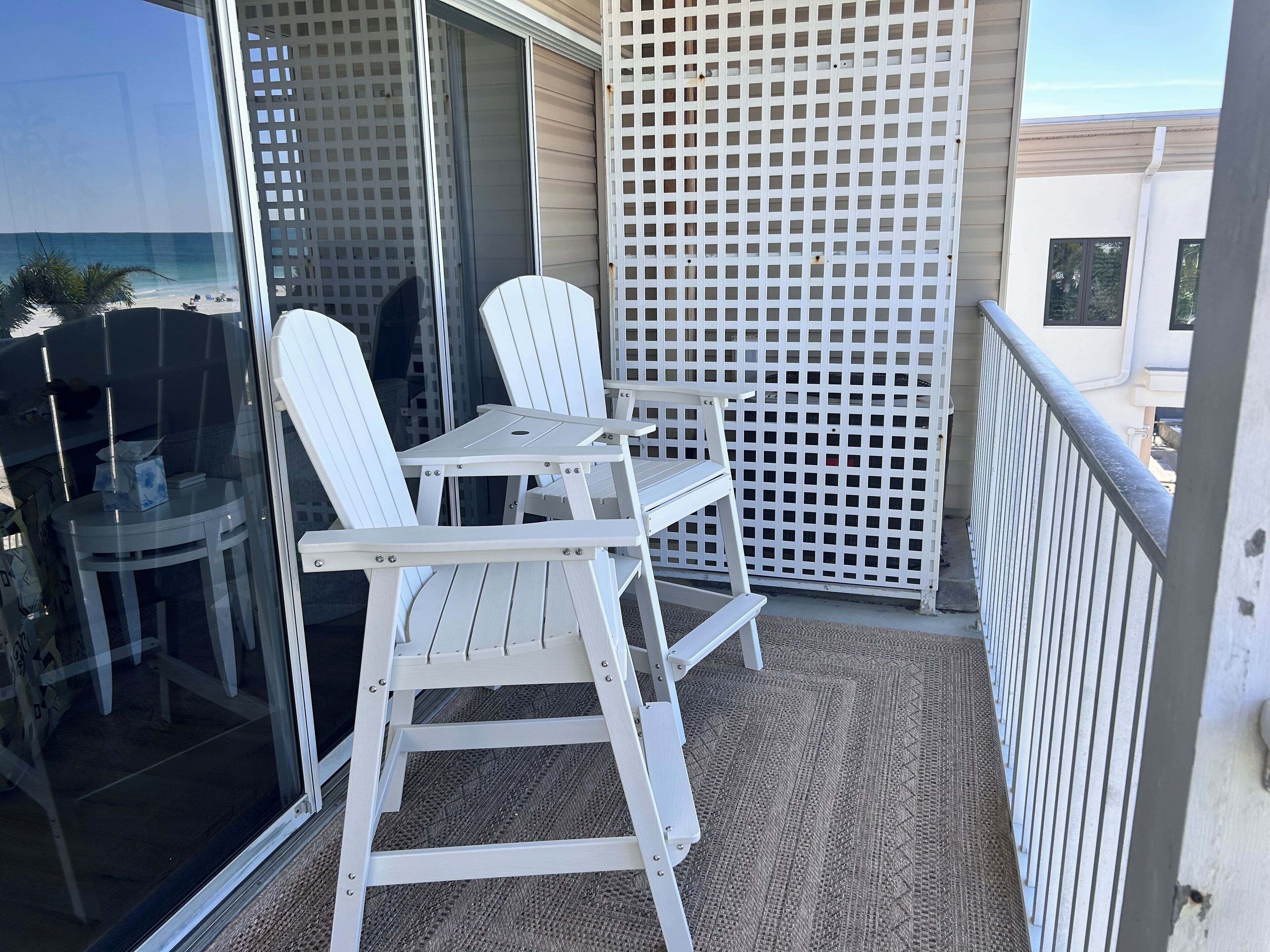 Un balcón con sillas blancas y una mesa, una puerta de vidrio y vista a la playa.