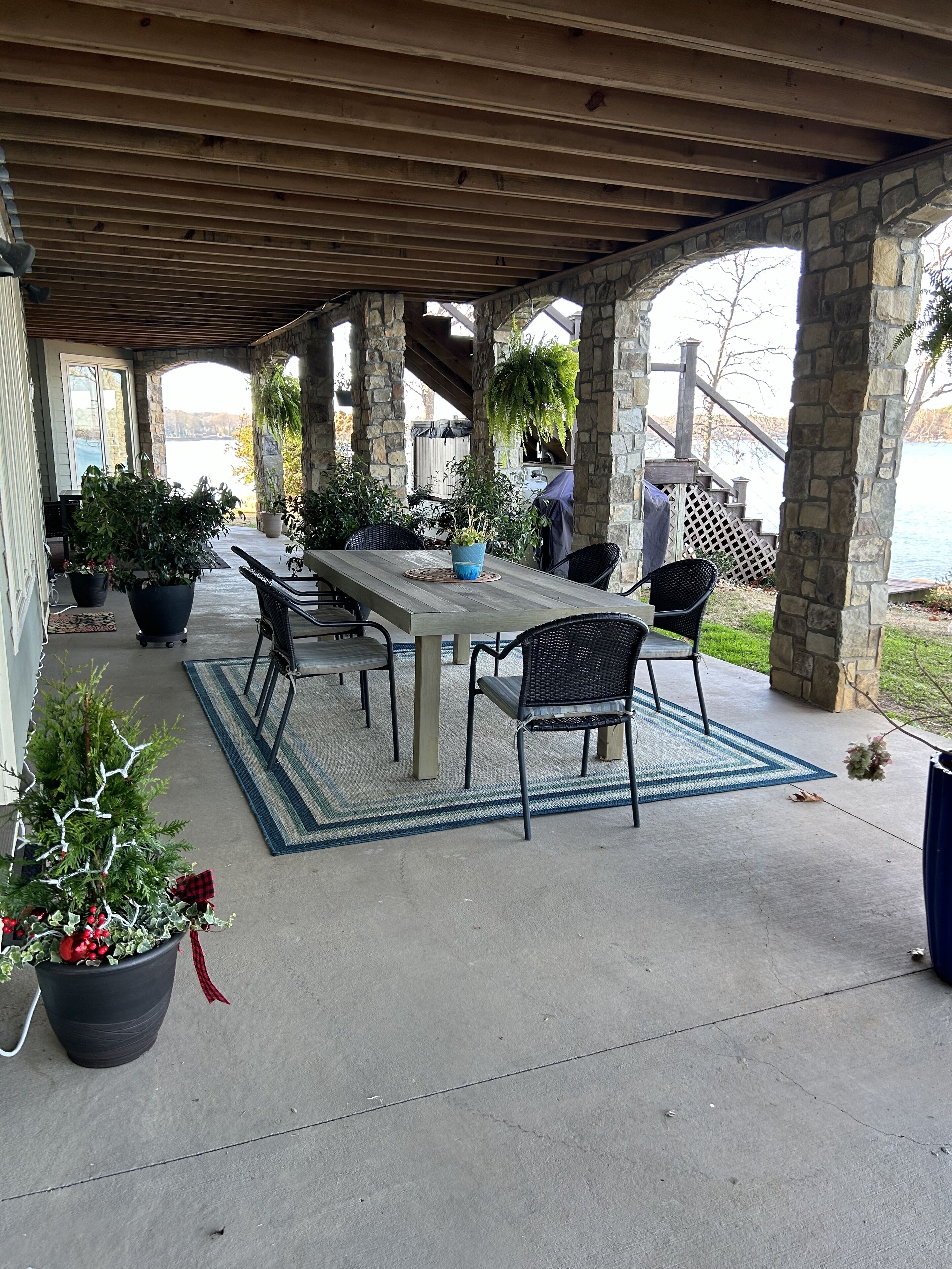 A covered patio area with a table and chairs, surrounded by potted plants and a view of a body of water.