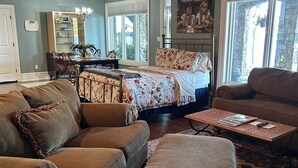 A cozy living room with a sofa, ottoman, and coffee table, a bed with floral bedding, a dining area with chairs and a table, and a large window with blinds.