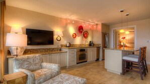 A modern kitchen with a flat-screen TV, a refrigerator, and a dining area with chairs. - Kuhio Shores 306:  Vacation On Kauai Oceanfront (Poipu)