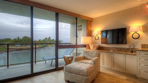 A spacious room with a large sliding glass door leading to a balcony overlooking a body of water. There is a comfortable chair and a small table on the balcony. The room has a flat-screen TV mounted on the wall, and there are two wall-mounted lamps. - Kuhio Shores 306:  Vacation On Kauai Oceanfront (Poipu)