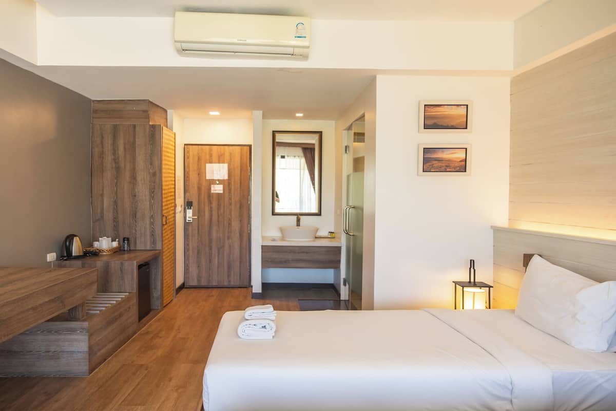 Ein Hotelzimmer mit Holztisch, einem Bett mit weißer Bettwäsche, einem Badezimmer mit Waschbecken und Spiegel sowie einer Wandklimaanlage.