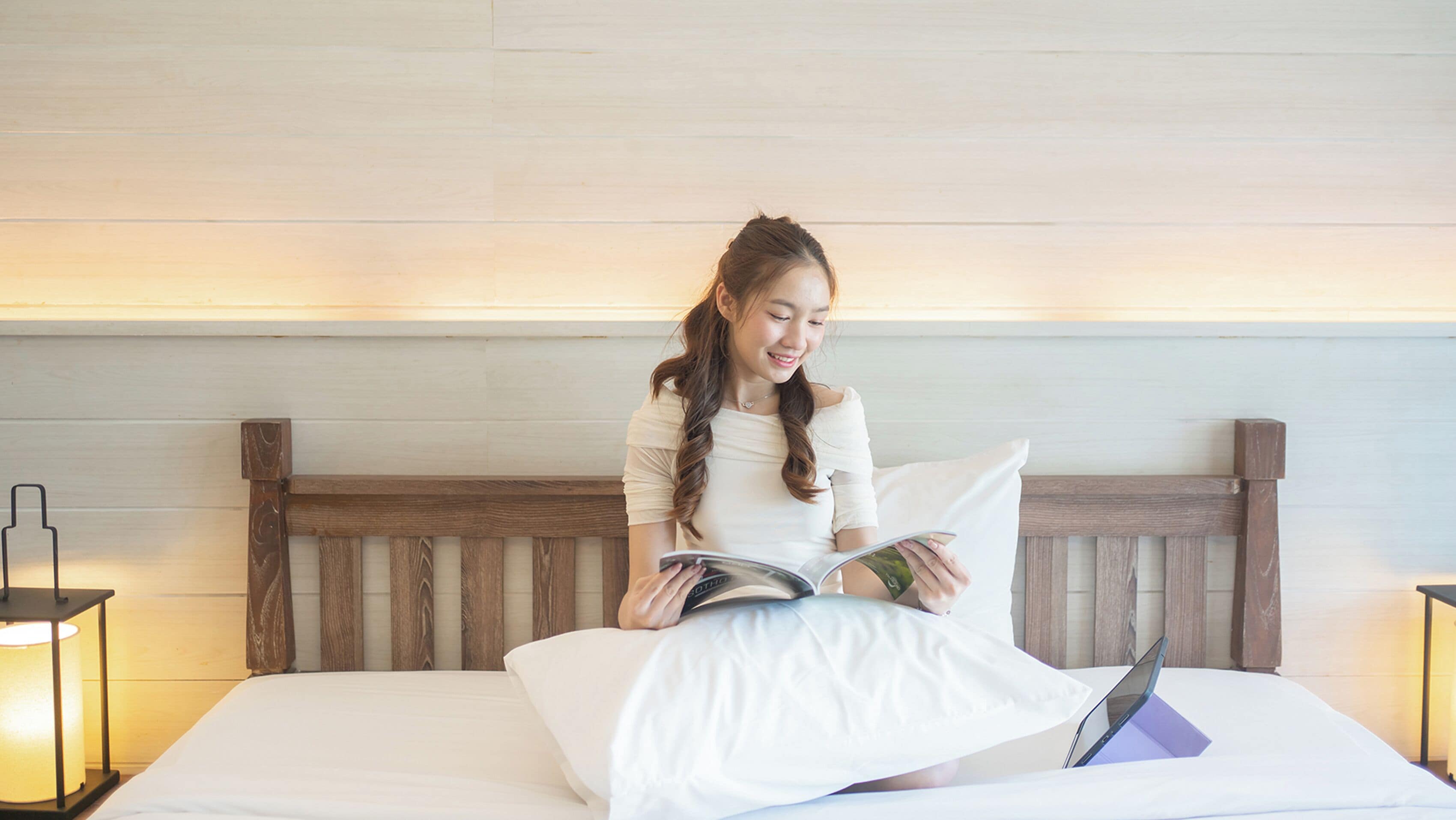 Une personne est assise sur un lit doté d’une tête de lit en bois, et lit un livre. Un ordinateur portable est posé sur le lit.
