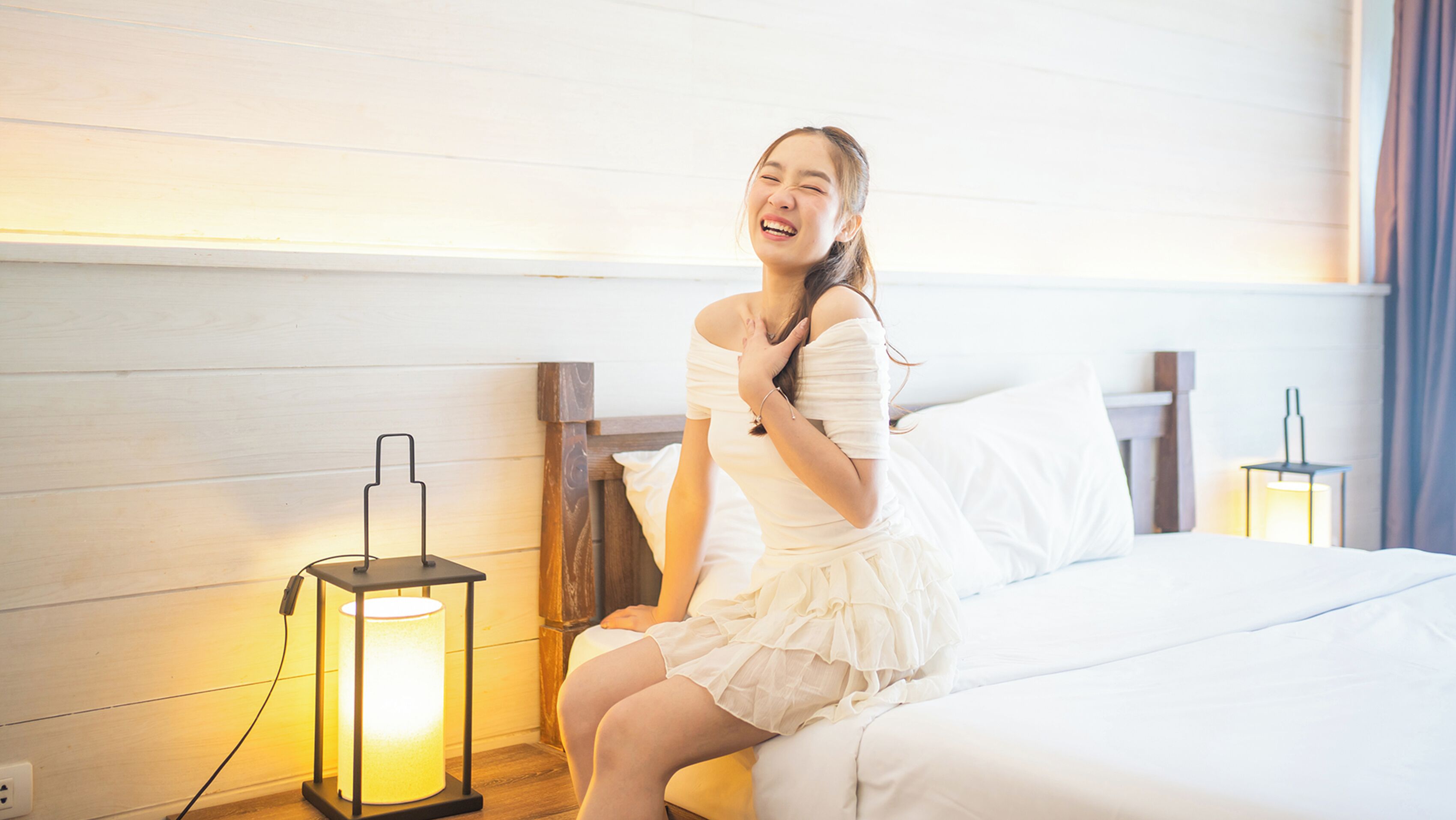 A person sitting on a bed in a room with wooden paneling, a nightstand with a lamp, and a wall-mounted light fixture.