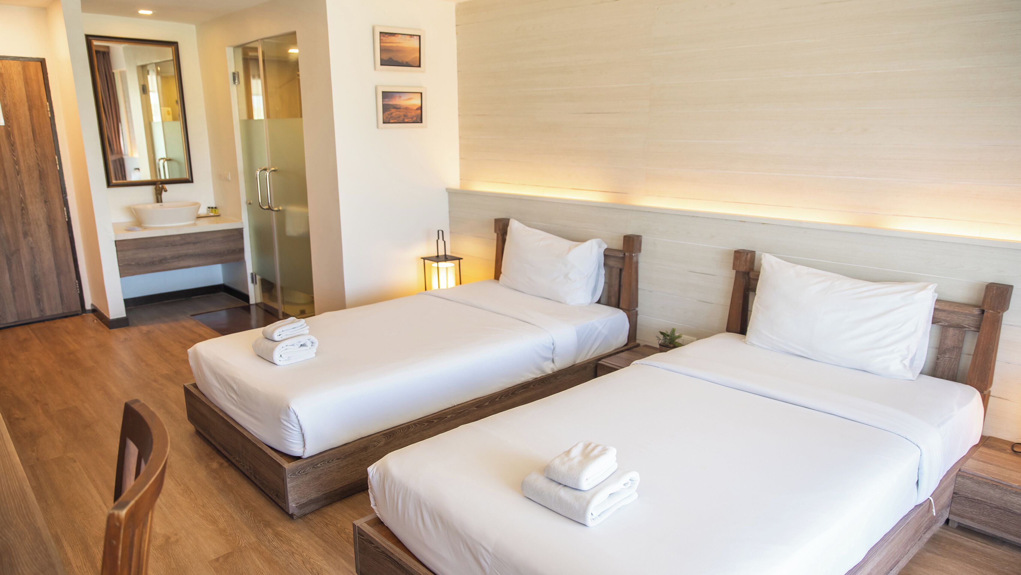 A hotel room with two beds, a wooden floor, a bathroom with a mirror, and a small plant on the bedside table.