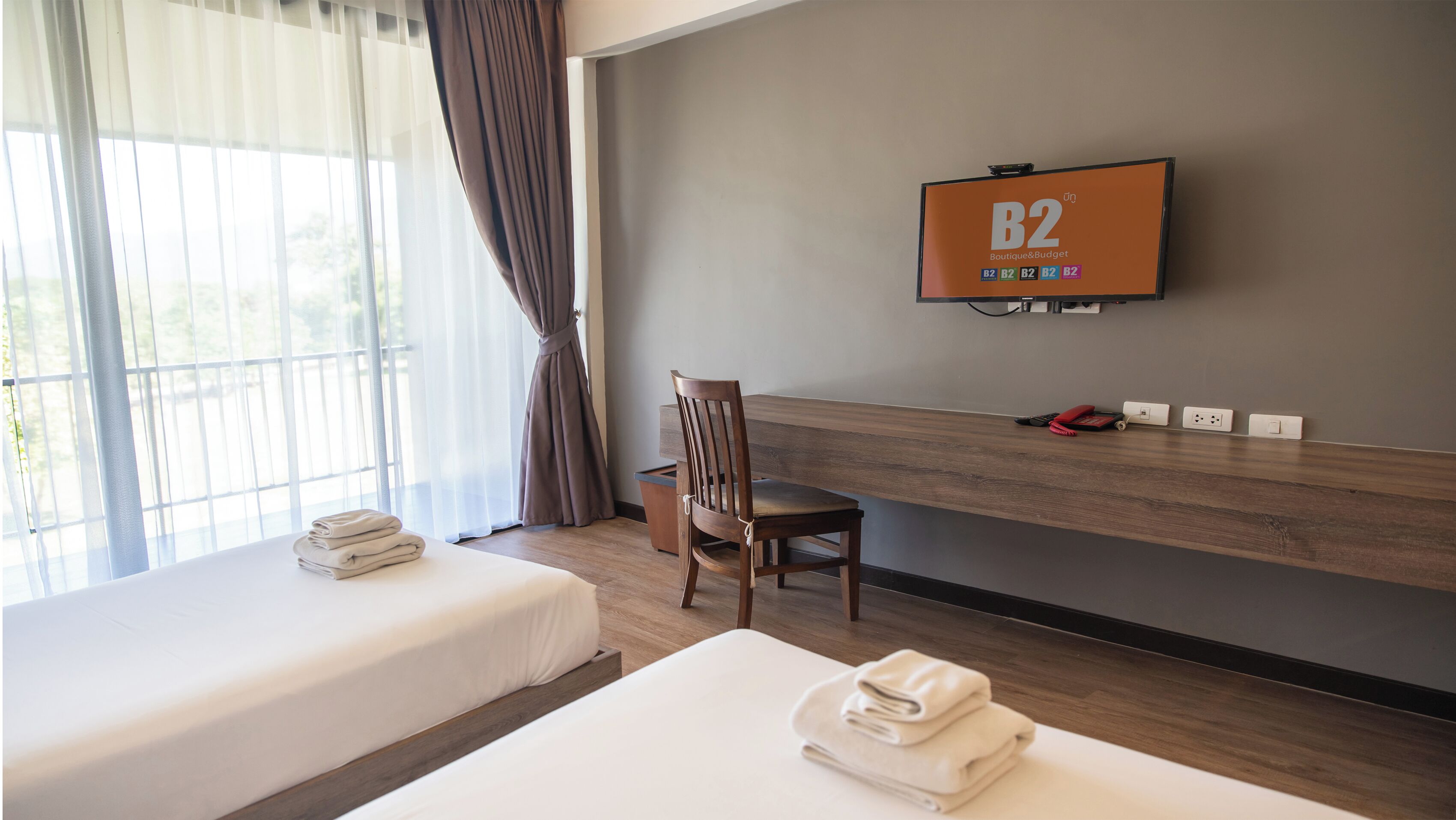 A hotel room with two beds, a TV on the wall, a desk with a chair, and a window with curtains.