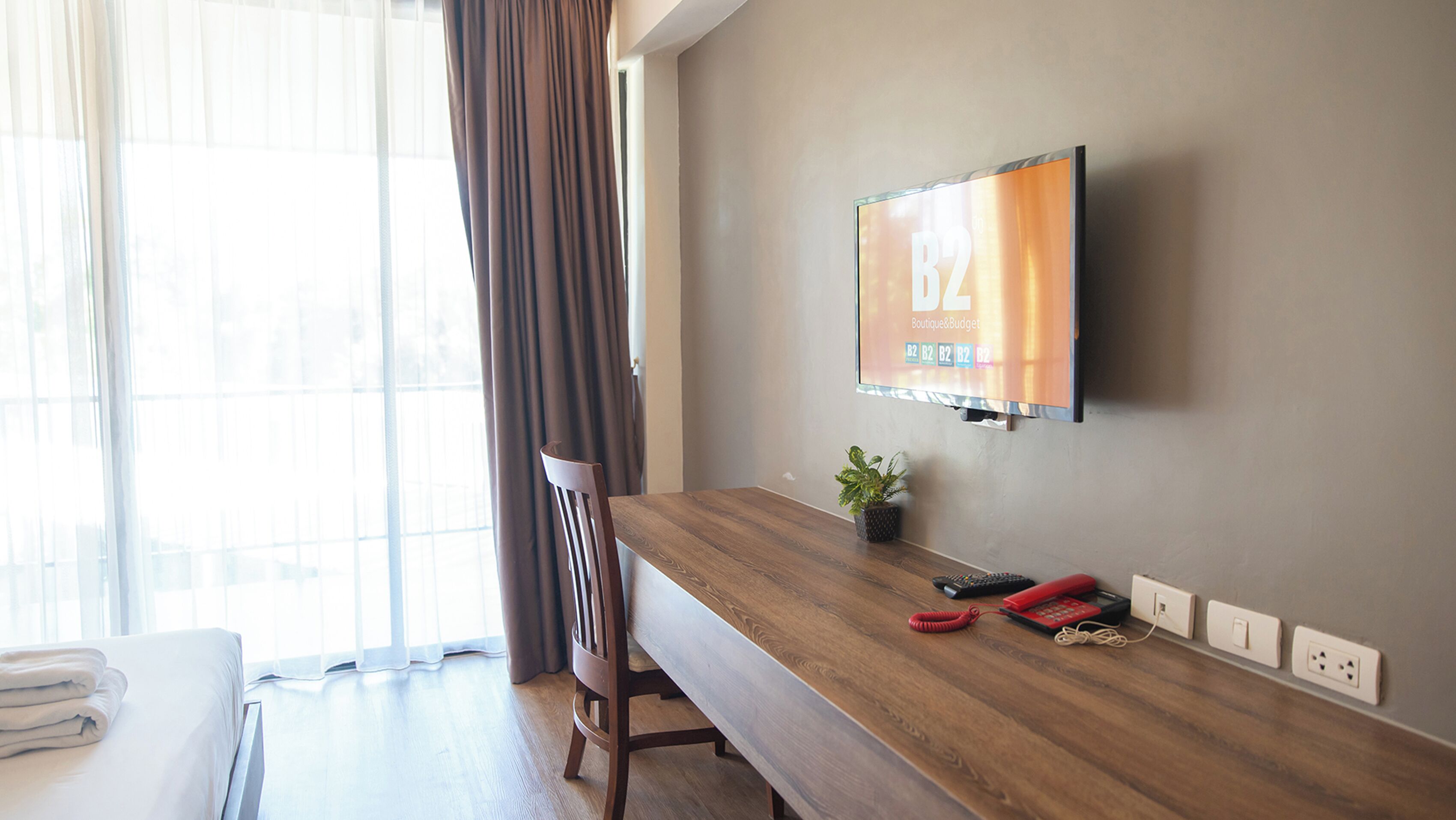 Hotellihuone, jossa on puinen työpöytä, seinälle asennettu televisio, tuoli, sänky ja ikkuna, jossa on verhot.