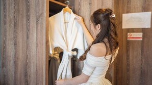 Uma pessoa de vestido branco está em um quarto de madeira com uma peça de roupa branca em um cabide.