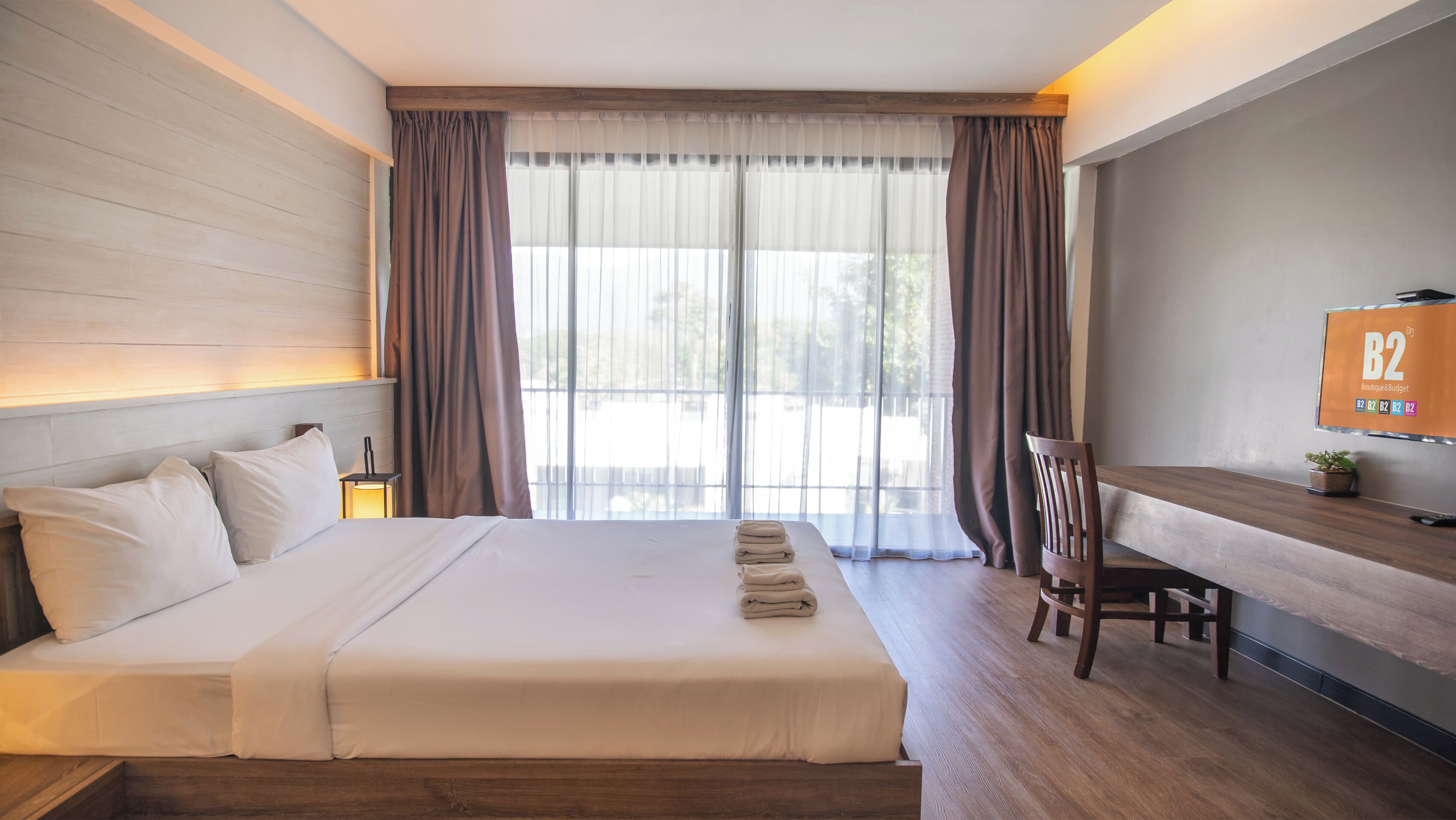 Una habitación de hotel con cama, escritorio, silla, televisor y ventana con cortinas.
