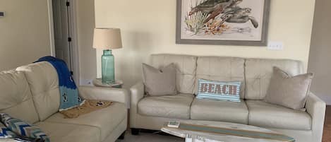 A living room with two beige sofas, a small round table, a lamp, and a framed picture of sea turtles on the wall.