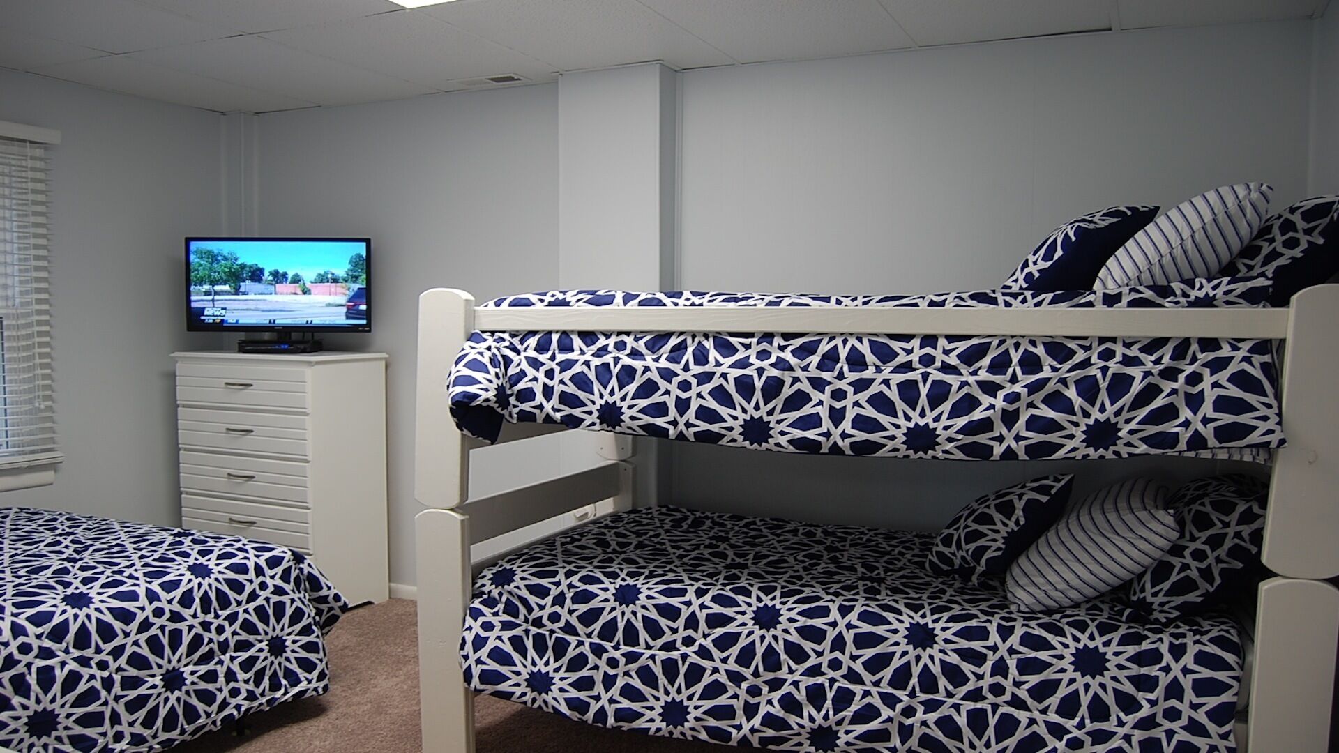 A bunk bed room with a television on a dresser, patterned bedding, and a window with blinds.