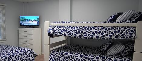 A bunk bed room with a television on a dresser, patterned bedding, and a window with blinds.