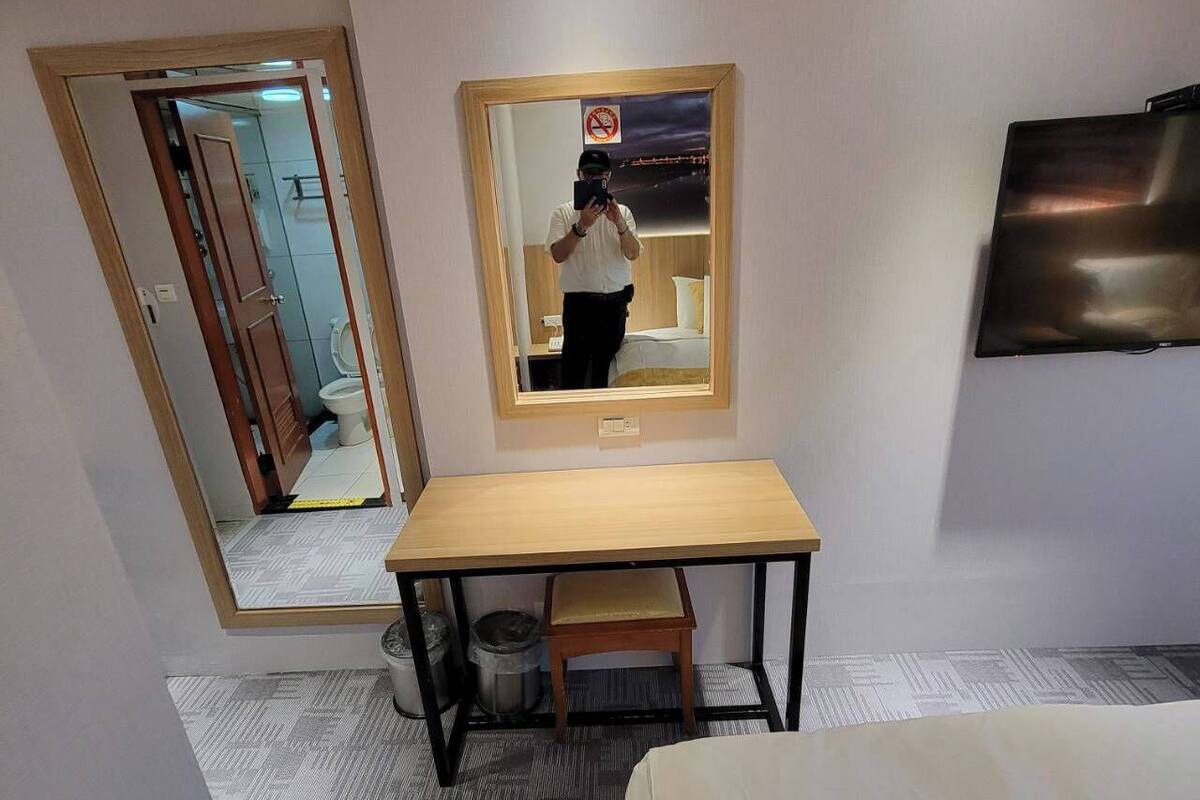 Una camera d'albergo con un letto, una scrivania, una sedia, un cestino, uno specchio e una televisione appesa al muro.