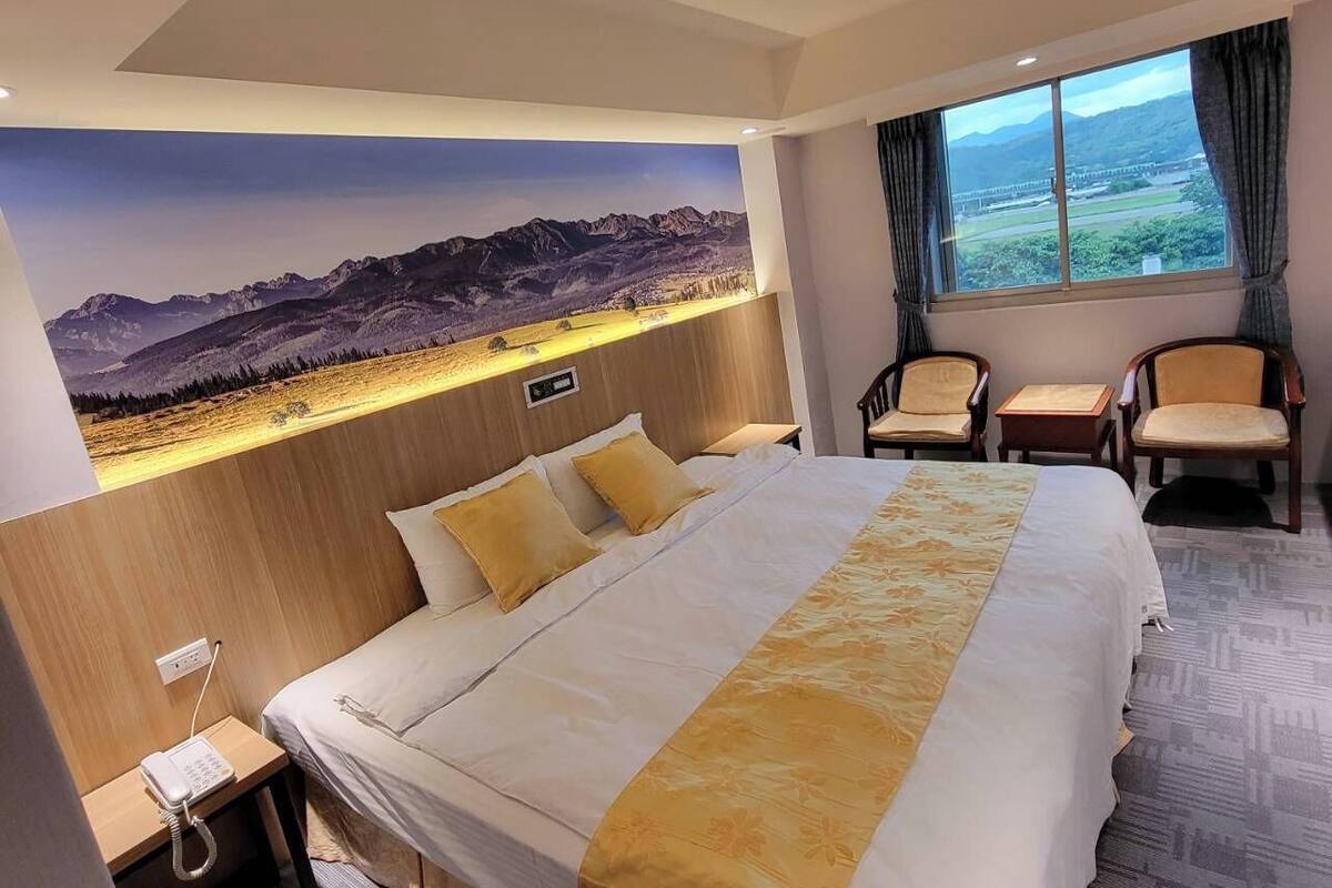 Camera d'albergo con un letto grande, due poltrone, un tavolino, una finestra vista montagne e un telefono fissato alla parete.