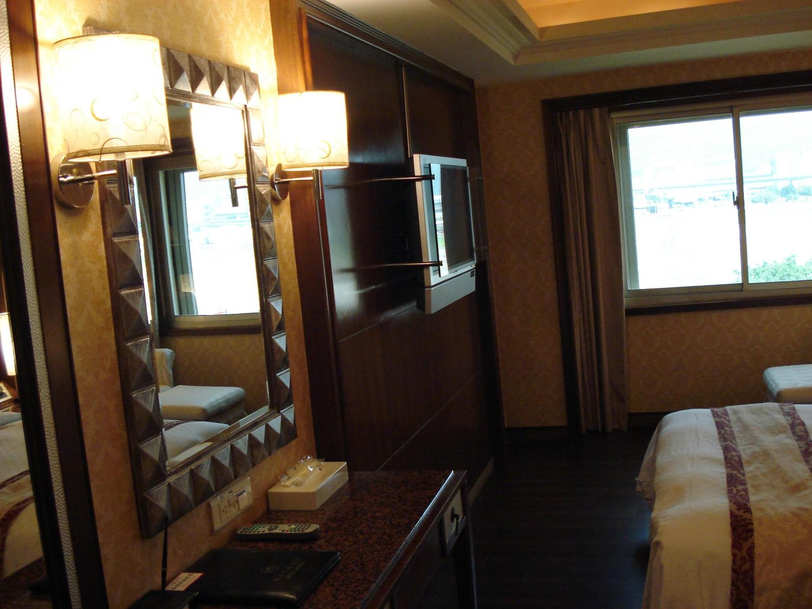 A hotel room with a bed, a TV mounted on the wall, a desk with a lamp, and a window with curtains.
