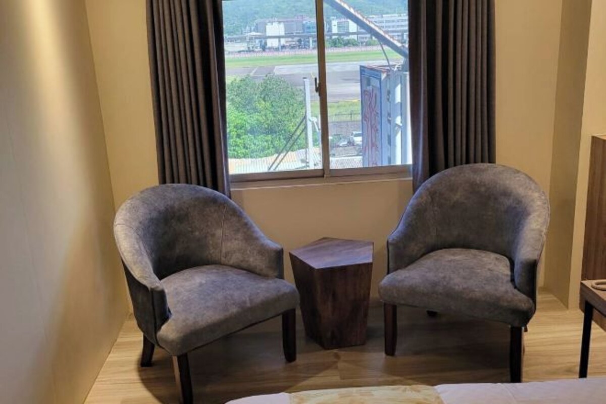 Une pièce avec deux fauteuils gris, une petite table d’appoint en bois et une vue sur un immeuble par une fenêtre ornée de rideaux foncés.