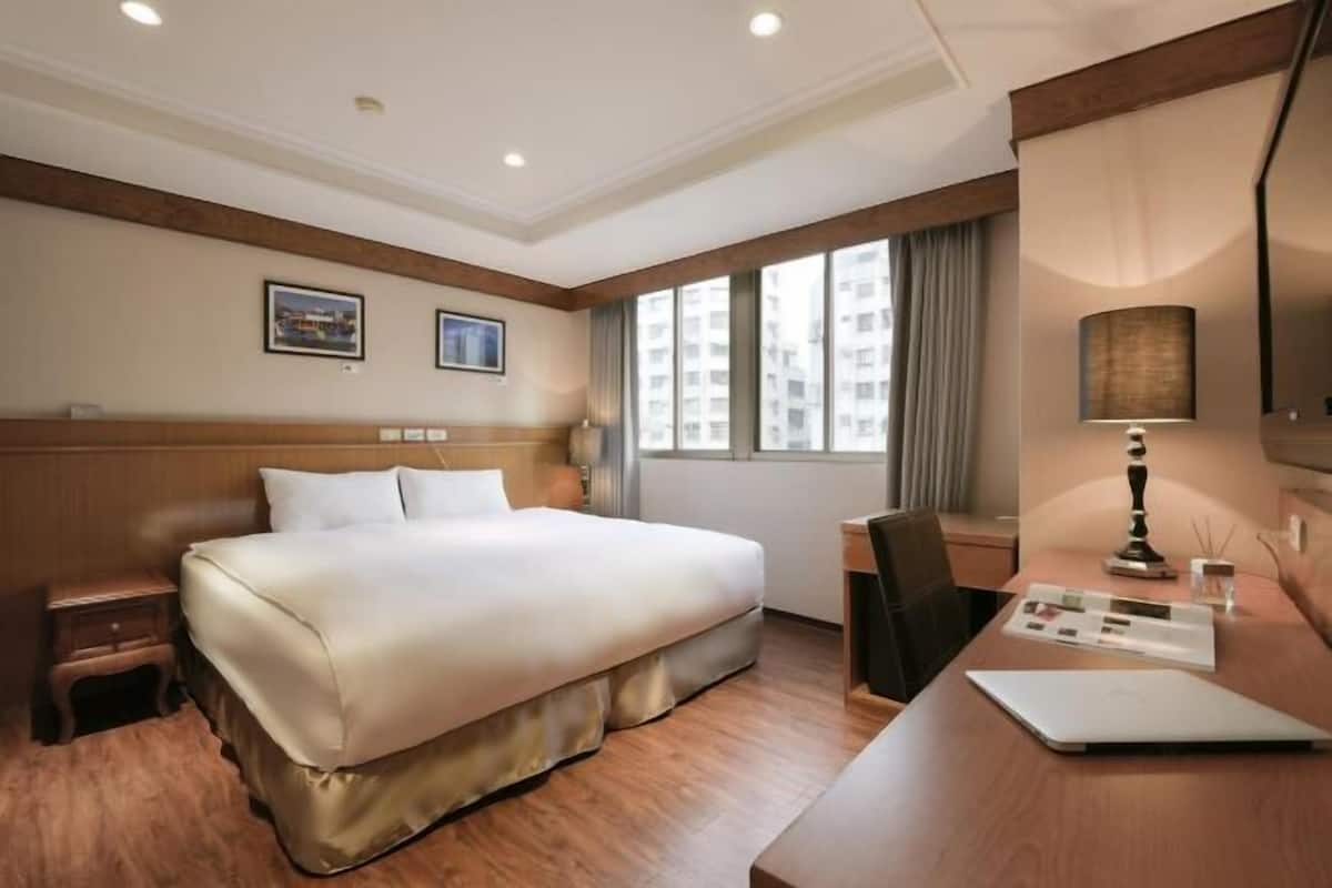 A hotel room with a large bed, a desk with a laptop, a chair, a nightstand, and a window with a view of buildings.
