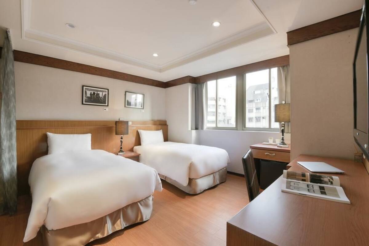 A hotel room with two beds, a desk, a chair, a lamp, and a window with a view of buildings.