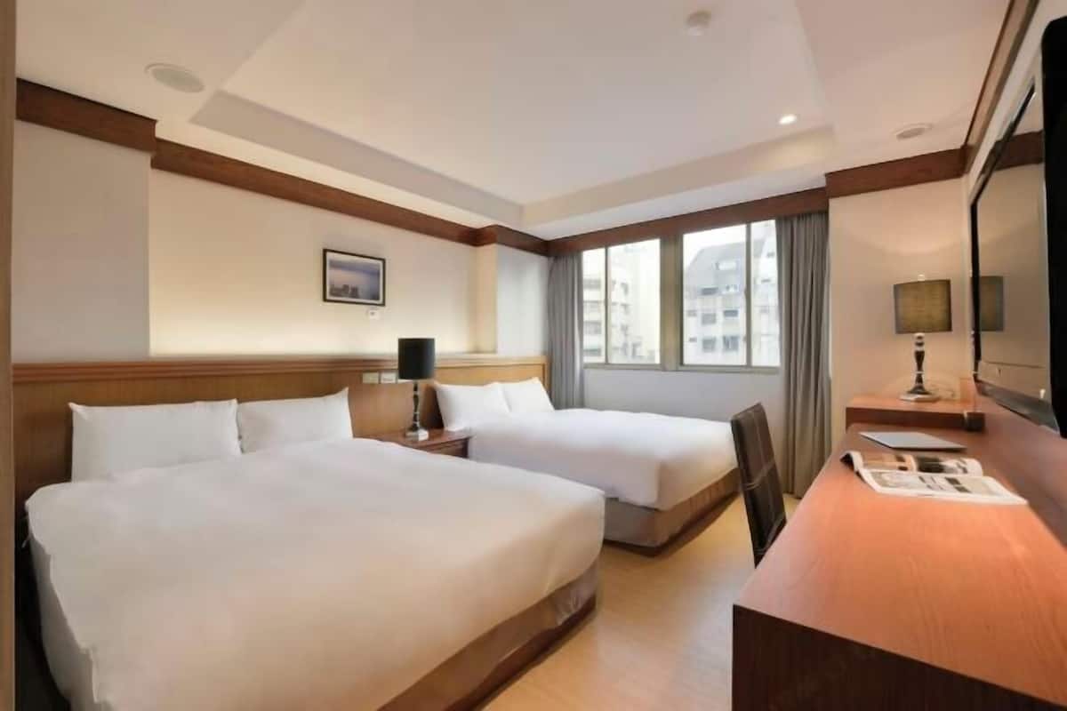 A hotel room with two beds, a desk, a chair, a lamp, and a window with a view of buildings.