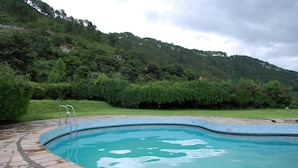 Une piscine bordée de pierres, avec un escalier en métal, entourée d’une végétation luxuriante et située au pied d’une colline.