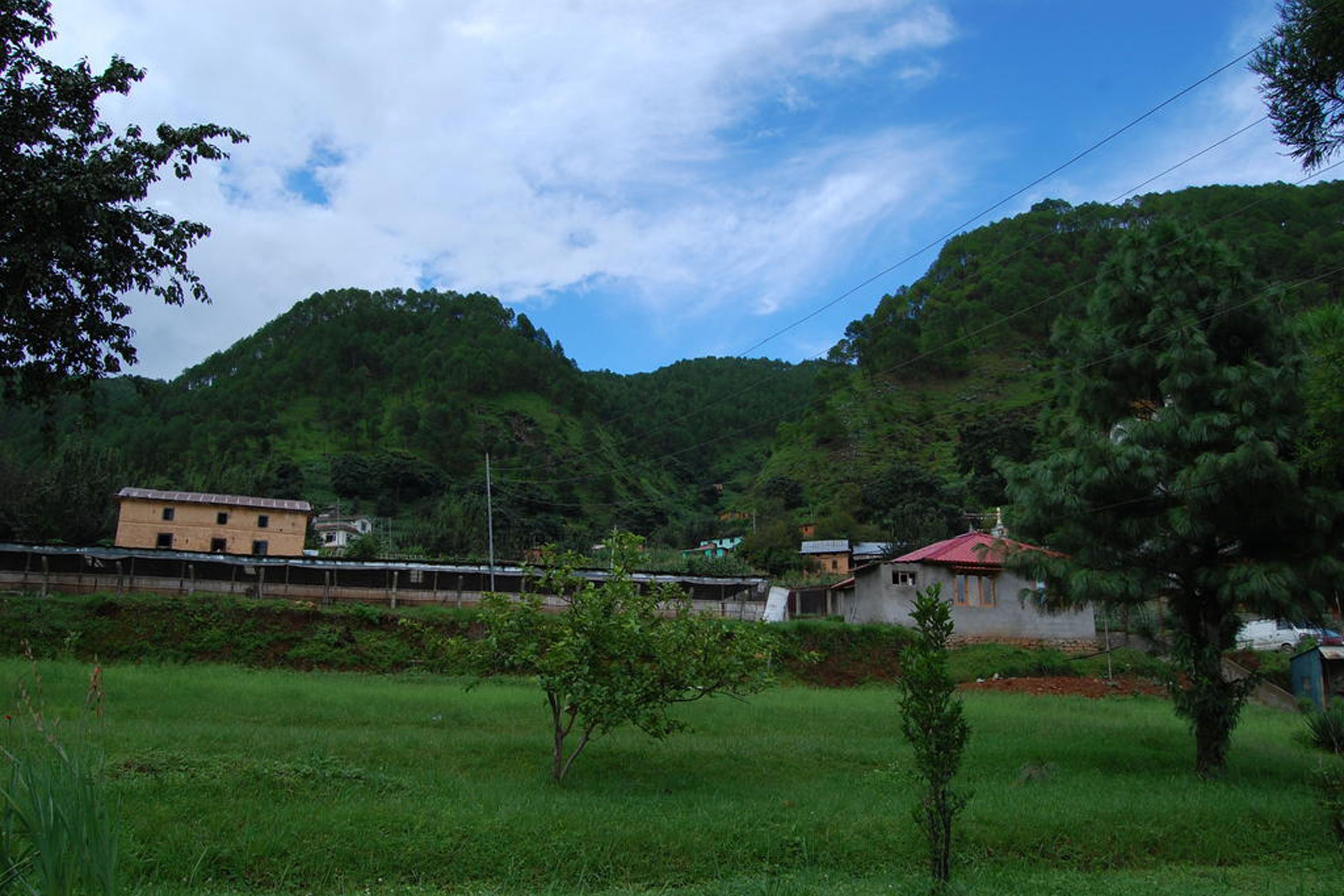 Paisaje rural con una construcción pequeña, una zona cercada y una colina al fondo.