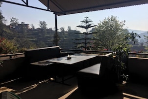 Una terraza con mesa y asientos, con vistas a un paisaje montañoso con árboles y edificios.