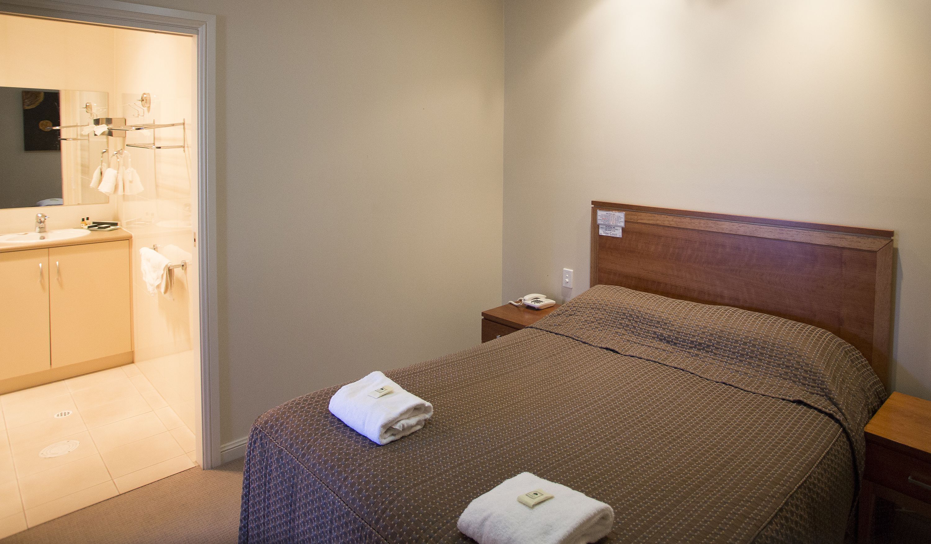 Habitación de hotel con cama, mesita de noche, teléfono y baño con lavabo y espejo.