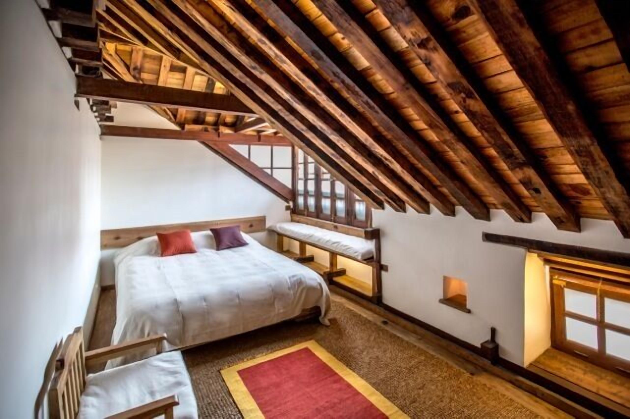 A bedroom with a sloped ceiling, a bed, a bench, and a window.