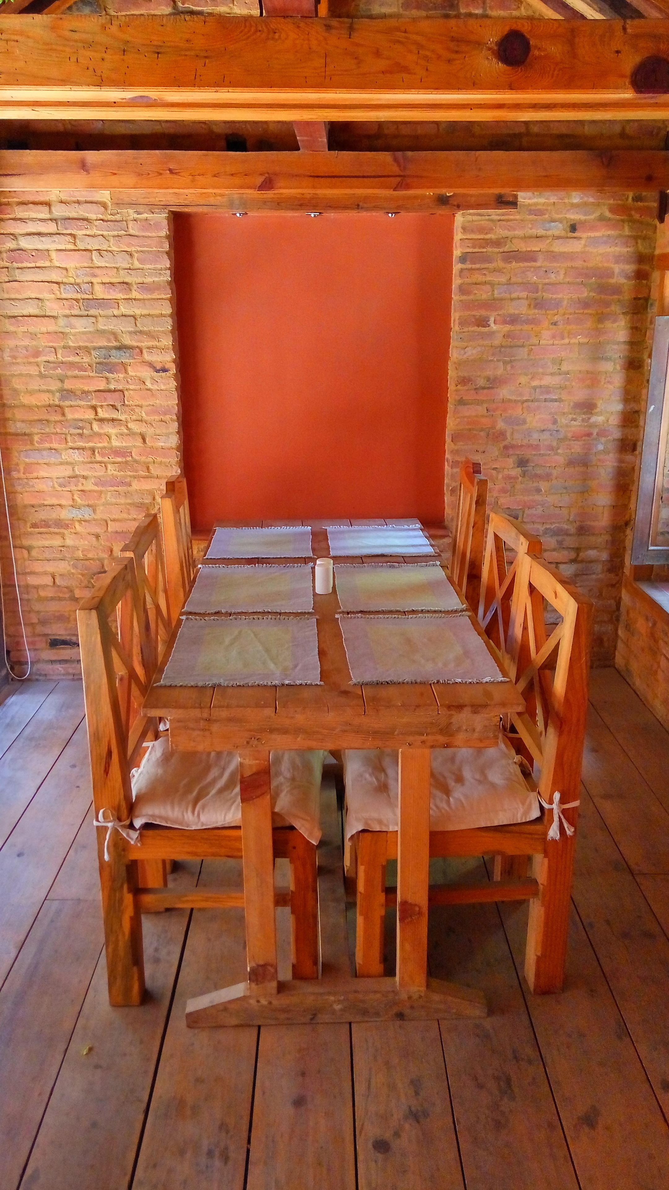 A wooden dining table with cushioned chairs, set against a brick wall and wooden ceiling beams.
