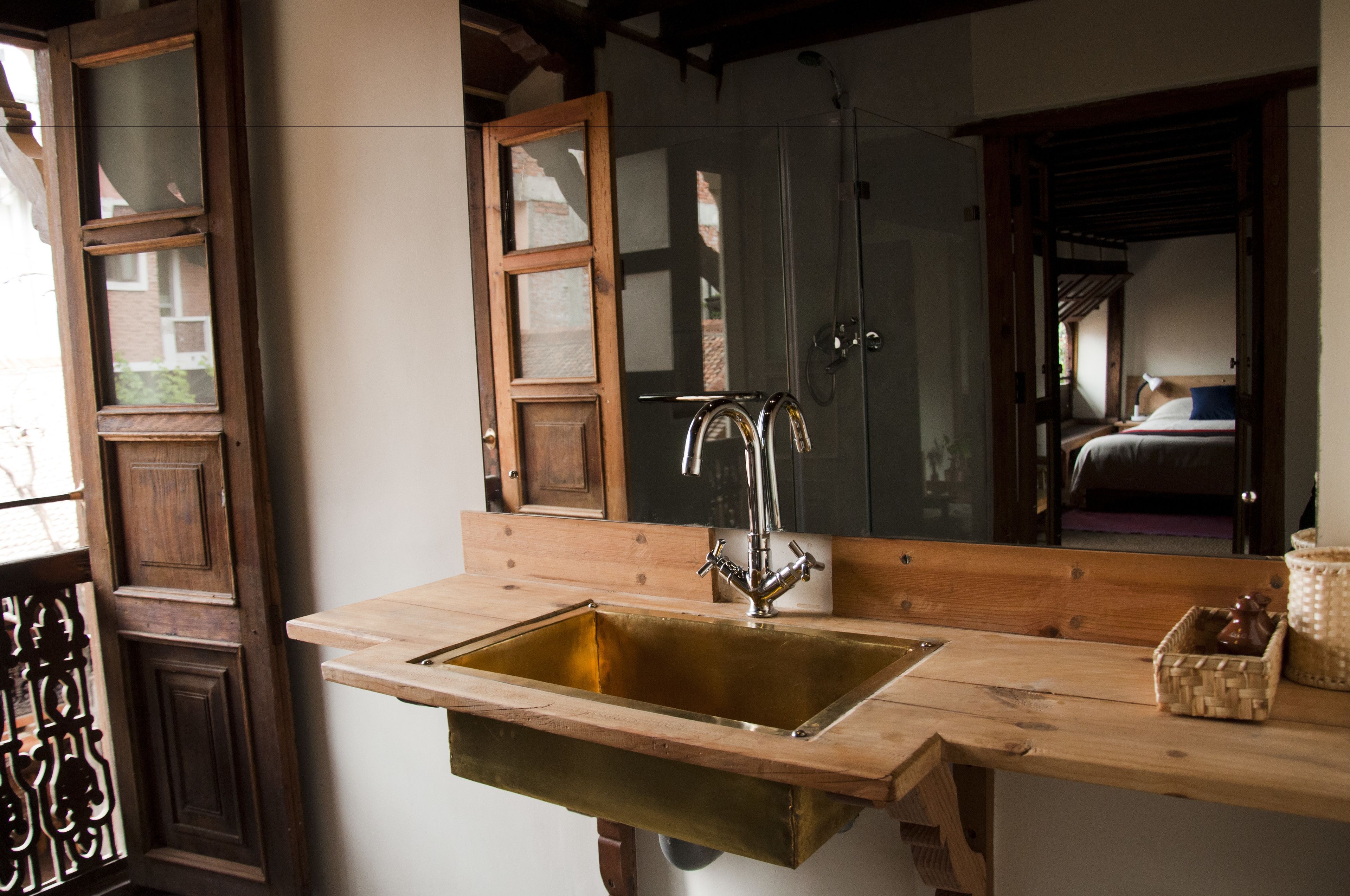 A bathroom with a wooden vanity, a brass sink, and a mirror reflecting a bedroom with a bed and a nightstand.