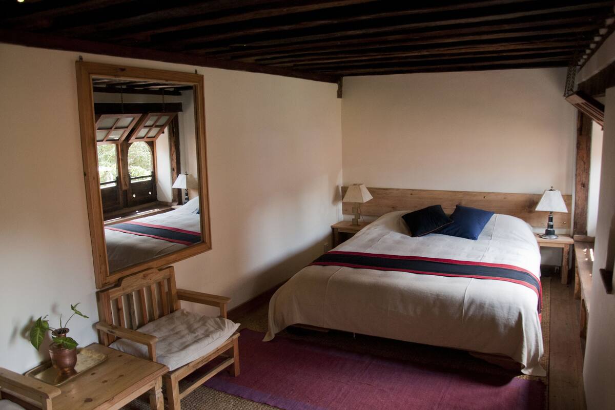 A bedroom with a large mirror, a bed with a striped blanket, a wooden nightstand, a bedside table, and a chair.