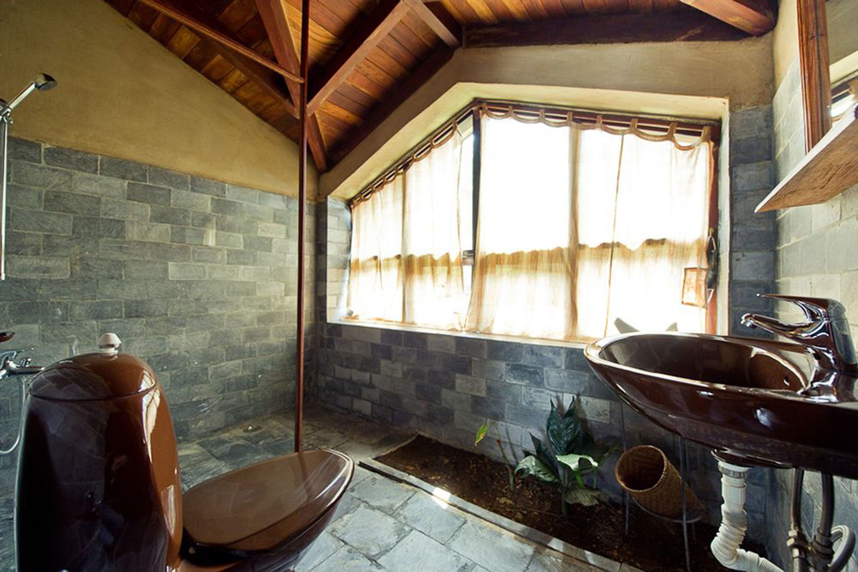 A bathroom with a stone wall, a large window with curtains, a wooden bench, and a sink.