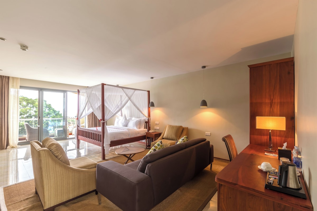 A hotel room with a four-poster bed, a sofa, a desk with a lamp, and a view of the outdoors.