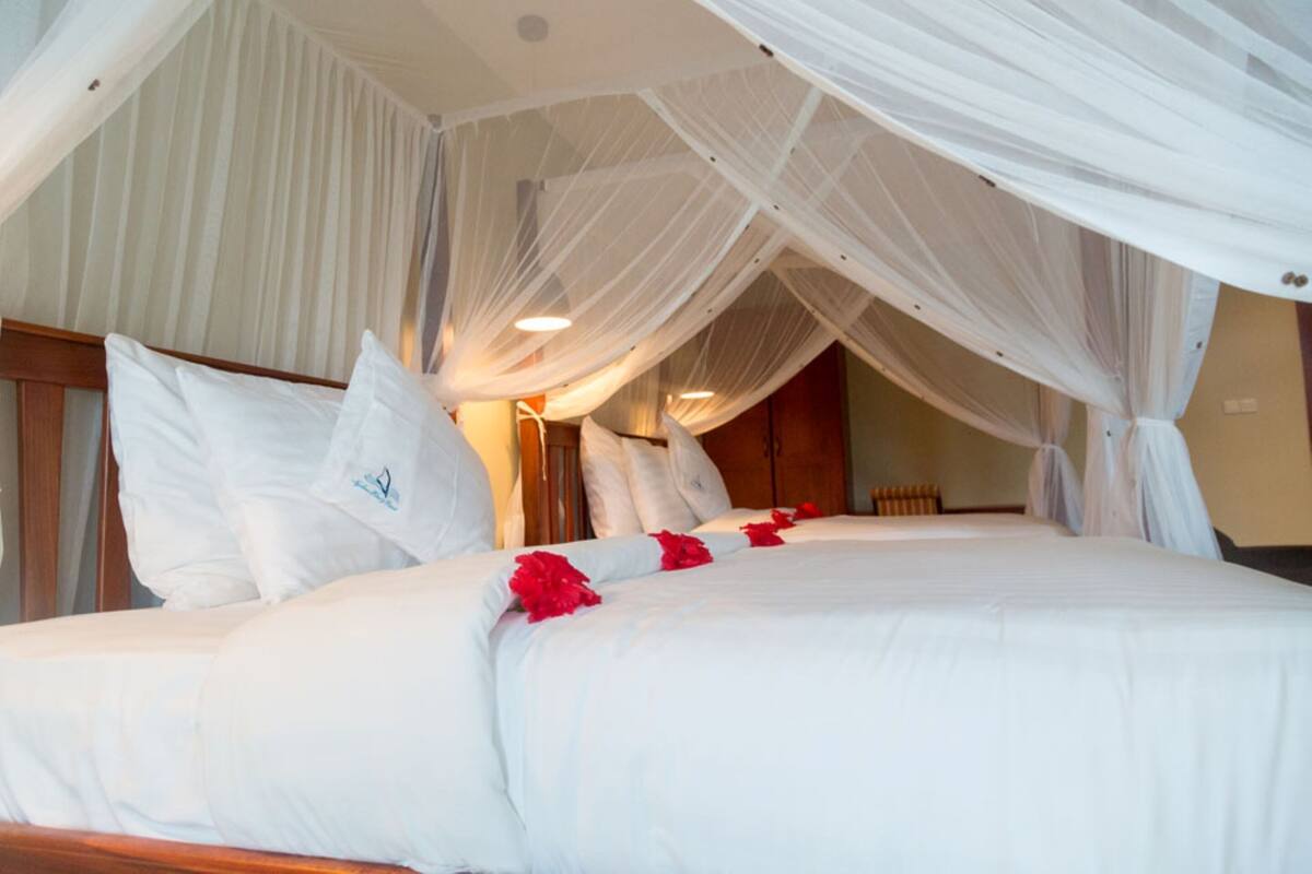 Cama com roupa de cama branca e flores vermelhas, cabeceira de madeira e cortinas brancas leves.