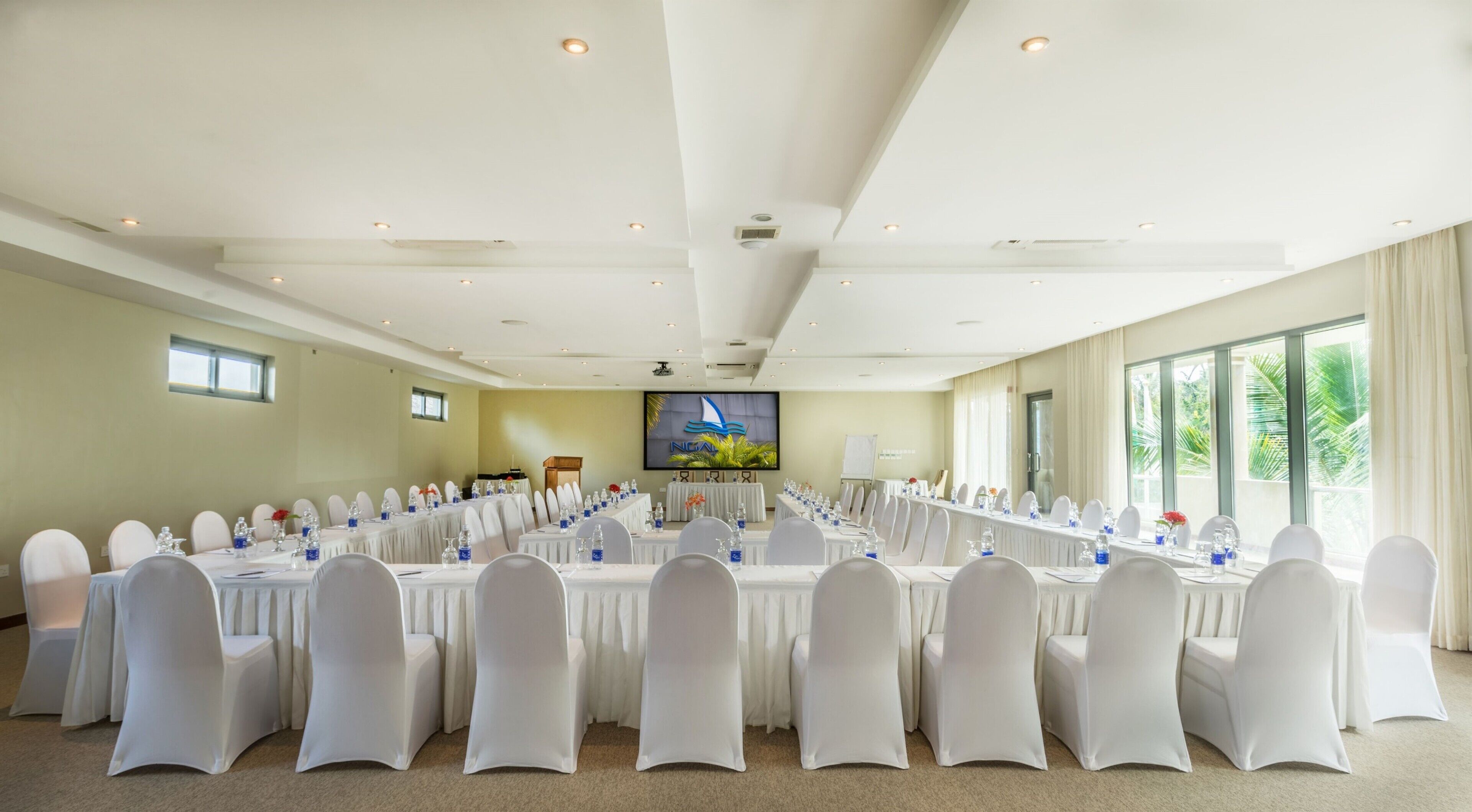 A large conference room with multiple tables and chairs, a large screen, and a view of palm trees through the windows.