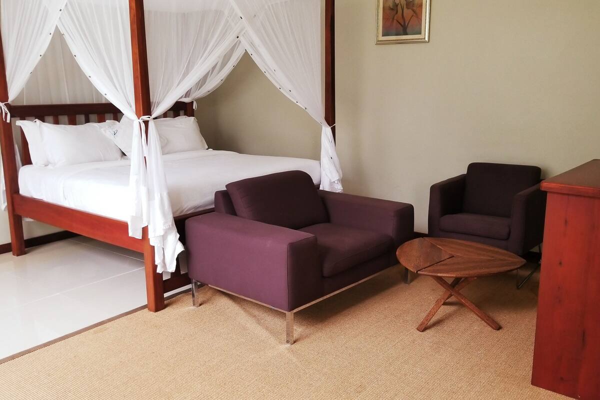 Uma cama de dossel com estrutura de madeira e roupa de cama branca, um sofá roxo, uma pequena mesa de madeira e uma poltrona escura em um quarto com parede lisa e um quadro.
