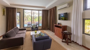 A modern living room with a sofa, armchair, and ottoman, a TV mounted on the wall, and a view of a pool outside.