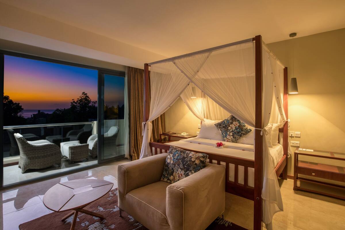 Quarto com cama de dossel, sofá, mesa de cabeceira e vista do pôr do sol por uma grande janela.