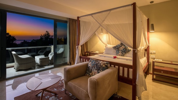 A bedroom with a four-poster bed, a sitting area with a chair and a small table, and a view of the sunset through a large window.